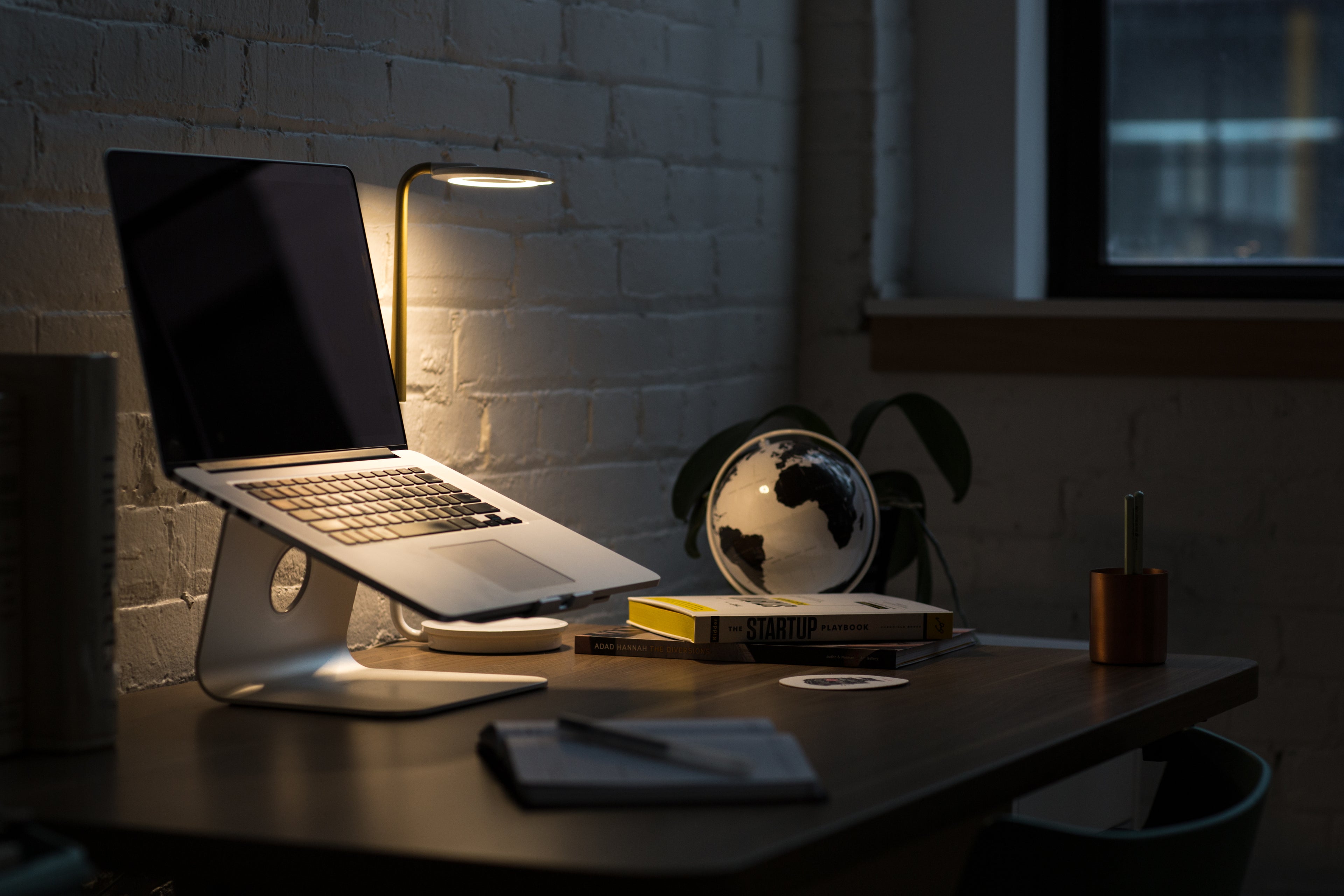 A laptop on a stand sits on a desk beside a lit lamp, books, a globe, a pen, and a notebook. The background features a white brick wall and a window, creating a cozy workspace atmosphere.